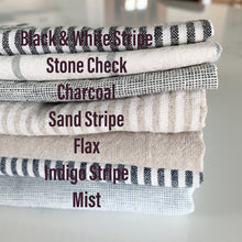 Load image into Gallery viewer, stack of folded linen showing color options for handmade dish drying mat including black and white stripe, stone check, charcoal, sand stripe, flax, indigo stripe, mist