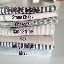 Load image into Gallery viewer, stack of folded linen showing color options for handmade dish drying mat including black and white stripe, stone check, charcoal, sand stripe, flax, indigo stripe, mist
